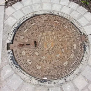 Centrální erbovní štít s vyobrazením pražského mostu a věží dominuje tomuto exempláři městské infrastruktury. Okolní geometrický vzor a nápisy jako 'PRAZSKA' a 'Kanalizace' jasně indikují jeho lokální příslušnost a funkční určení. Jedná se o robustní litinový artefakt, klasifikovaný pro vysoké zatížení D400, což svědčí o jeho odolnosti v urbanistickém prostředí.