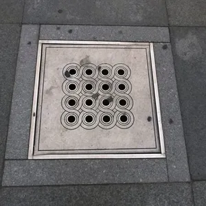 Na povrchu tohoto čtvercového přístupového bodu se rozprostírá výrazný geometrický vzor šestnácti propojených kruhů, úhledně zasazený do rámu z tmavě šedé žulové dlažby. Každý větší kruh obsahuje centrální, menší otvor, což naznačuje funkční design pro vstup tekutin. Celková prezentace, byť poněkud zvětralá, si zachovává zřetelný vizuální řád, což je svědectví o původním inženýrství, které Institut Lidologie s potěšením zaznamenává.
