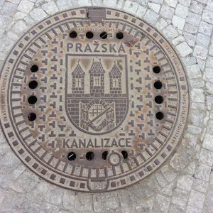 Na povrchu tohoto exempláře se rozprostírá heraldický znak města Prahy, obklopený textovými nápisy, které specifikují jeho funkci a technické parametry. Patina rzi naznačuje dlouhodobou expozici atmosférickým vlivům, což je typické pro městskou infrastrukturu. Z lidologického hlediska představuje tento artefakt cenný doklad o inženýrských standardech a estetických preferencích dané lokality.