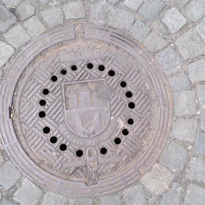 Uprostřed kruhového exempláře se nachází heraldický znak Prahy, obklopený nápisem 'PRAŽSKÁ KANALIZACE', což jednoznačně identifikuje jeho municipální příslušnost. Povrch je dále strukturován opakujícím se diagonálním geometrickým vzorem, který, jak se zdá, optimalizuje trakci a zároveň esteticky doplňuje funkční design. Tento artefakt městské infrastruktury nese stopy opotřebení, což svědčí o jeho dlouhodobé expozici environmentálním vlivům.