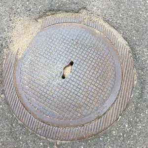 Hrubý mřížkovaný vzor pokrývá centrální část tohoto kulatého litinového poklopu, zatímco jeho okraj zdobí svislé, paralelní žebra, která se ztrácejí v okolním asfaltu. Povrch nese známky rzi a opotřebení, s nepravidelným otvorem pro klíč, který narušuje jinak přísnou geometrii. Kolem dokola se hromadí písek a drobné kamínky, které se mísí s drsnou texturou silnice.